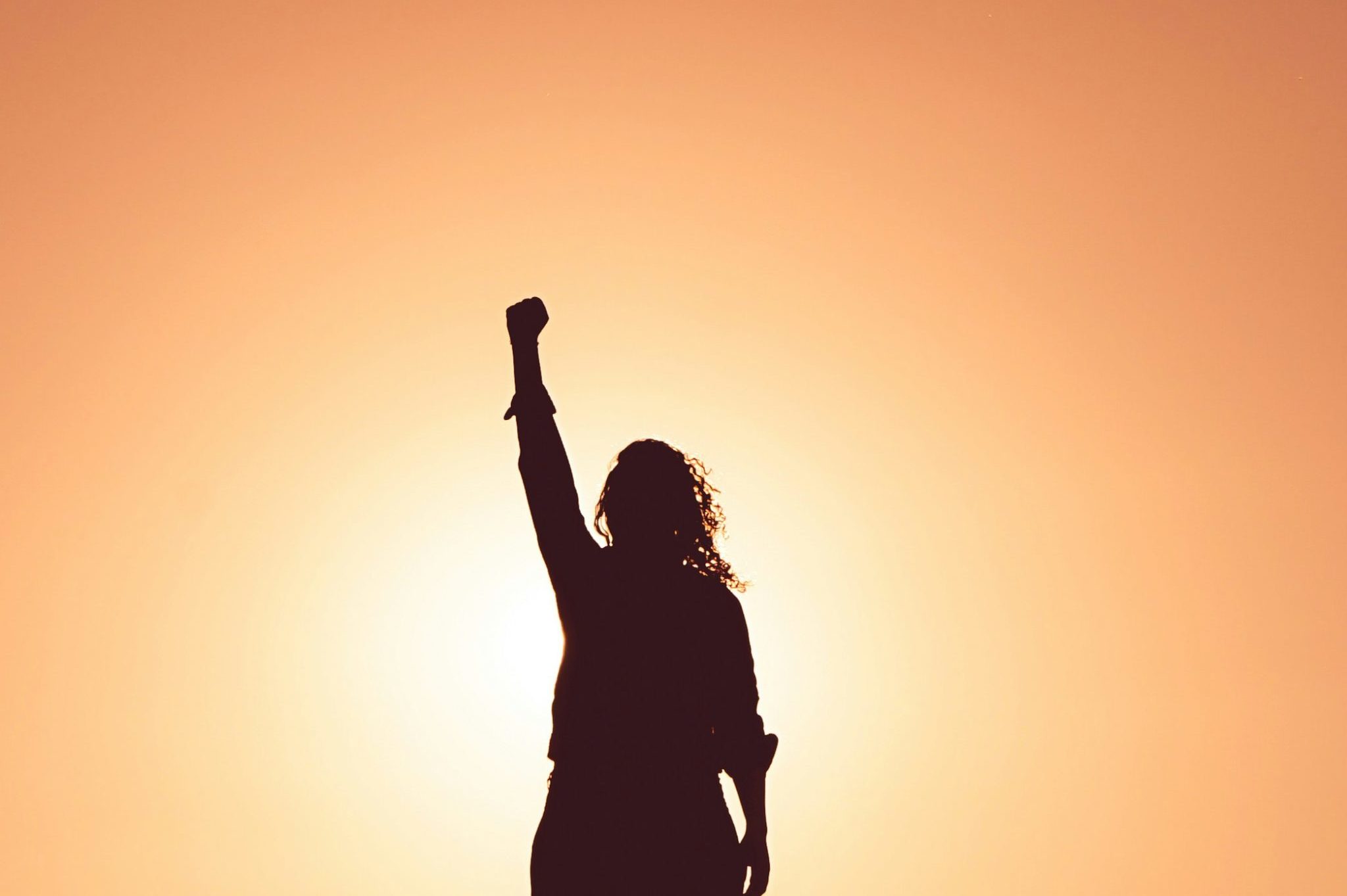 miguel-bruna-TzVN0xQhWaQ-unsplash Silhouette de femme en position de vainqueure, le poing levé, sur fond de coucher de soleil.